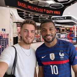 Two men smiling for a selfie inside a cosmetics store. The man on the right wears a blue and red France national football team jersey with the number 10, featuring the Nike swoosh and the French Football Association crest.