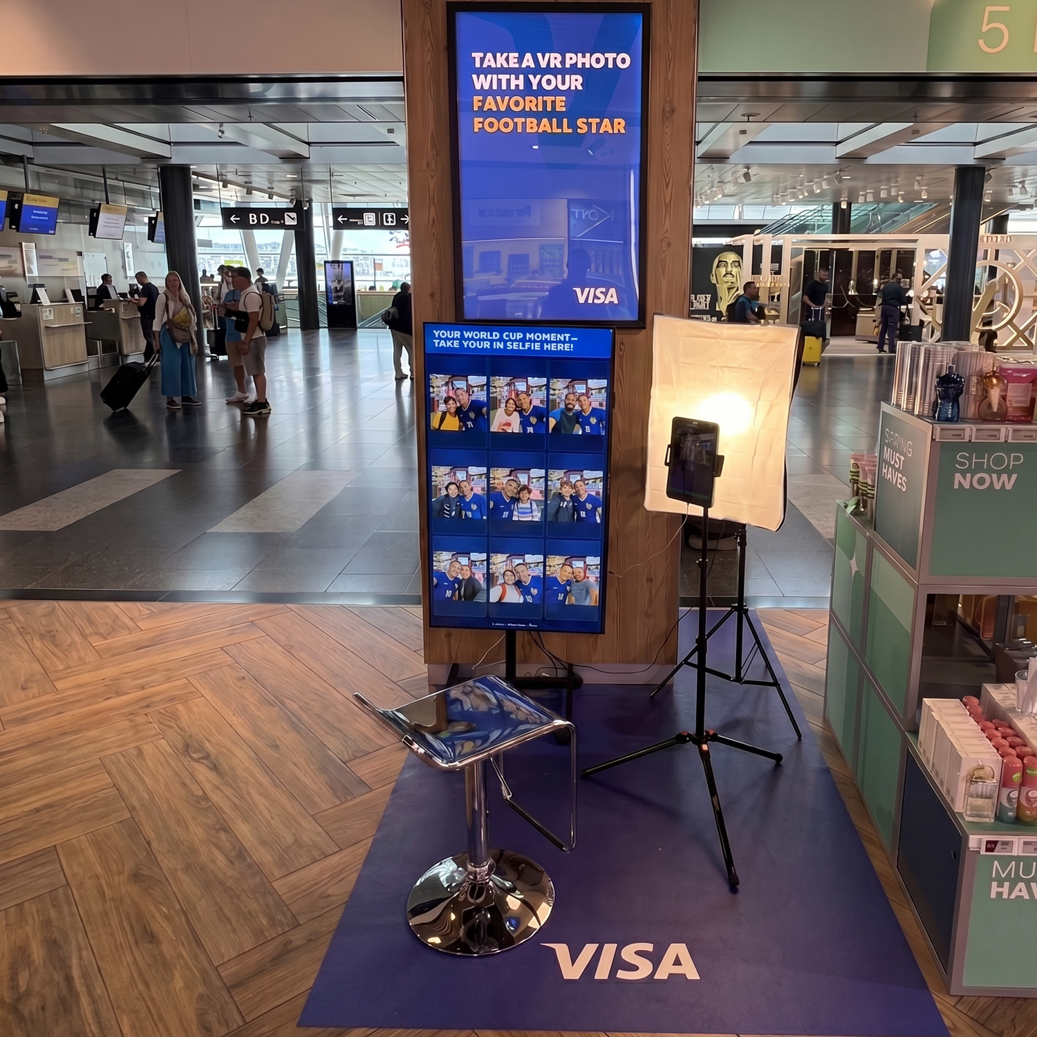 A VISA-branded photo booth setup in an airport or transit terminal, featuring a blue sign, sample photo thumbnails, a tripod-mounted light panel, a small stool, and a display stand on a blue floor mat.