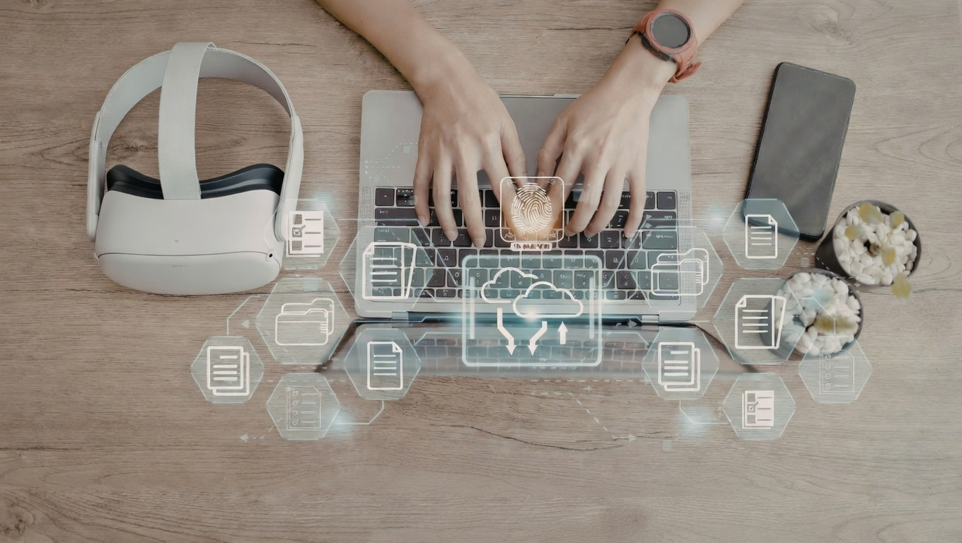 Person using a laptop with a VR headset and smartphone on a wooden table.