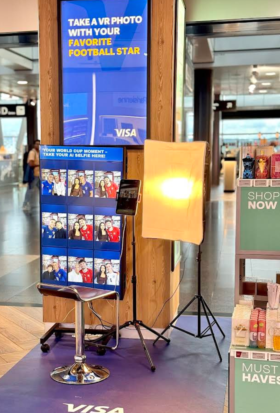 A VR photo booth setup in a retail environment, featuring a large screen showing sample photos and text, a stool, a softbox light, and a tablet on a tripod.