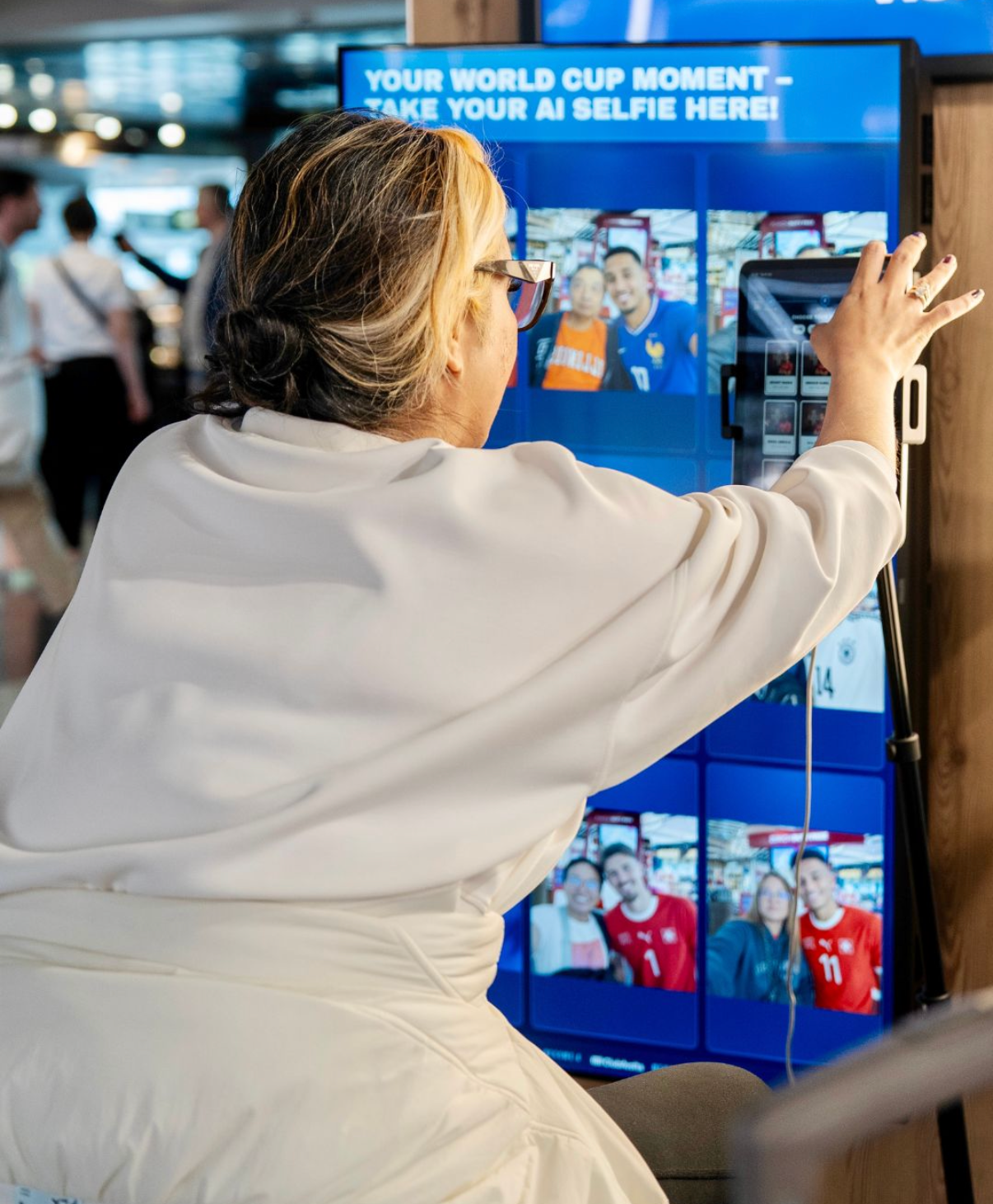 A person in a white robe stands in front of a large interactive digital kiosk screen, touching the display. The screen shows multiple video call thumbnails and the text 'YOUR WORLD CUP MOMENT - TAKE YOUR AI SELFIE HERE!'.